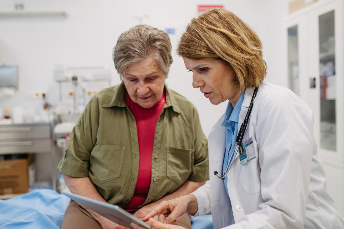2 ladies in a hospital. 1 is a doctor and another an elderly patient. The doctor is showing the patient something on an iPad.